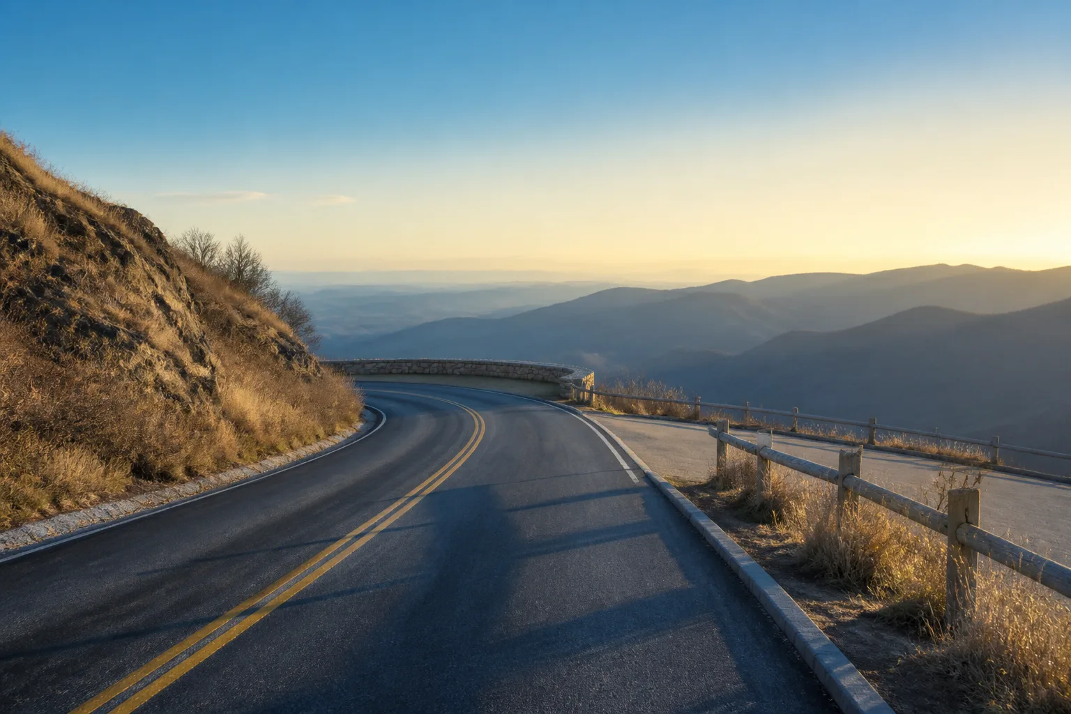 Empty two-lane highway at dawn curving toward a stone overlook, pale gold light on dry grass, a lone roadside pulloff with a weathered rail, long shadows across the asphalt, cool blue remaining in the far ridges, 1920x1080