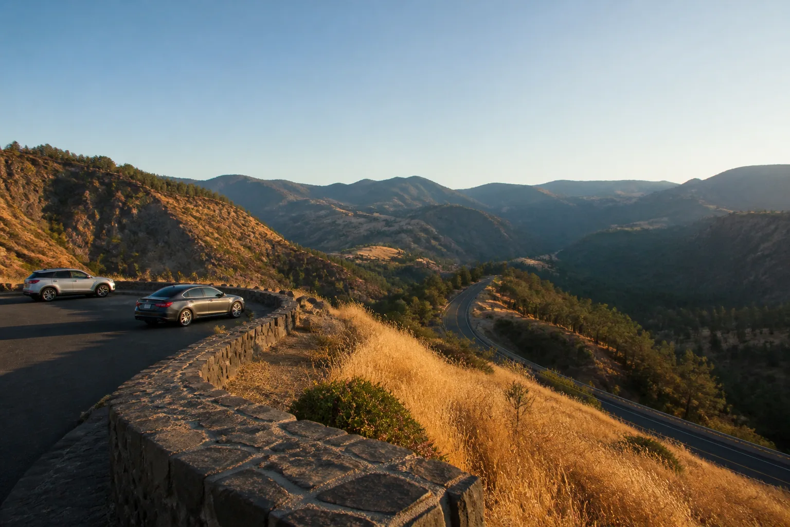 Stone overlook rail above a winding highway, small shared parking bay with two cars spaced apart, dry grass in late afternoon light, distant ridges under a clear sky, 1600x900