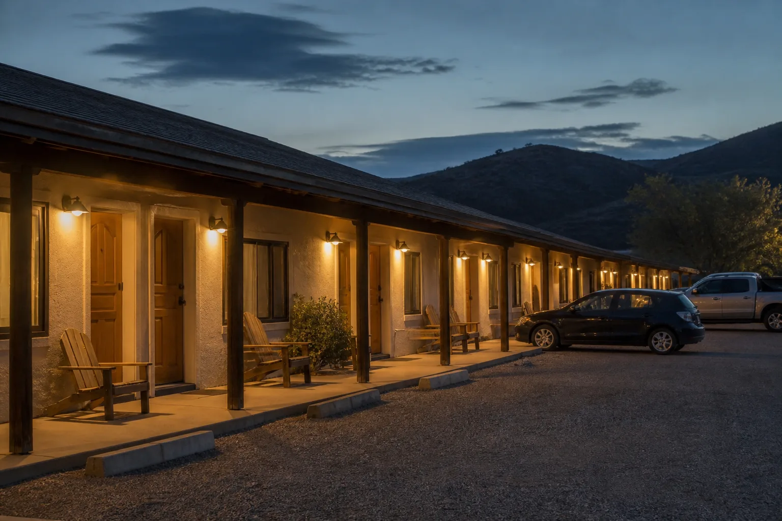 Single-story roadside motel at dusk, warm porch lights over numbered doors, a few parked cars in a gravel lot, empty wooden chairs, dim hills behind the roof line, 1600x900
